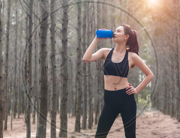 Beauty Asian Sport Woman Resting And Holding Drinking Water Bottle And Relaxing In Middle Of Forest After Tired From Jogging. Girl Looking Attraction View. Workout Concept. Lifestyle Theme