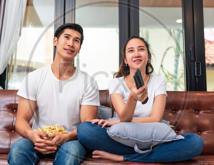 Asian Couples Watching Television Together On Sofa In Their Home. People And Lifestyles Concept. Vacation And Holiday Concept. Honeymoon And Pre Wedding Theme. Happy Family Activity In Valentines Day