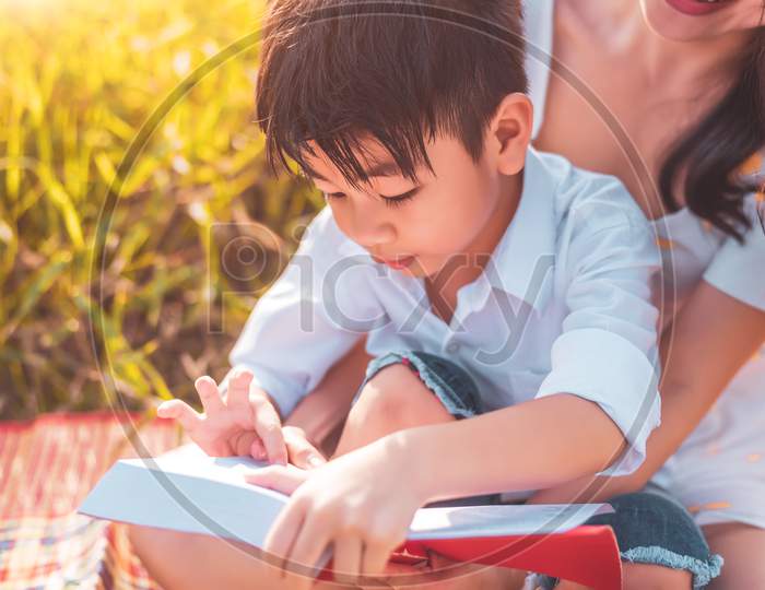 Little Asian Boy And His Mother Reading Tale Story Books At Meadow Field. Mother And Son Learning Together. Celebrating In Mother Day And Appreciating Concept. Summer People And Lifestyle Education