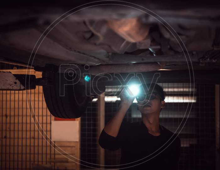 Lonely Asian Male Shining Flashlight To Examine Car Under Chassis Of Automotive Vehicle At Old Abandoned Garage In Night Scene. Haunted And Horror. Fantasy And Mystery Concept. Halloween Theme.