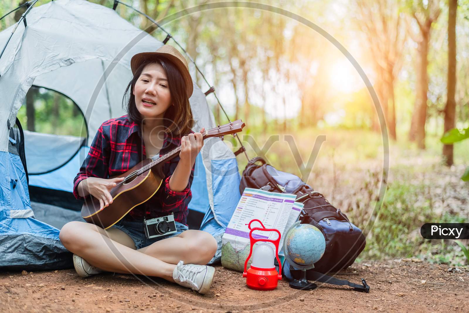 Beautiful Asian Woman Playing Ukulele In Front Of Camping Tent In Pine Woods. People And Lifestyles Concept. Adventure And Travel Theme.
