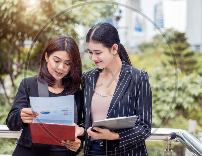 Two Young Asian Businesswomen Looking Into Document File Folder For Analyzing Profit Or Sale Break Even Point After Marketing. Business Teamwork Employees Of Lifestyle Working Women Concept.