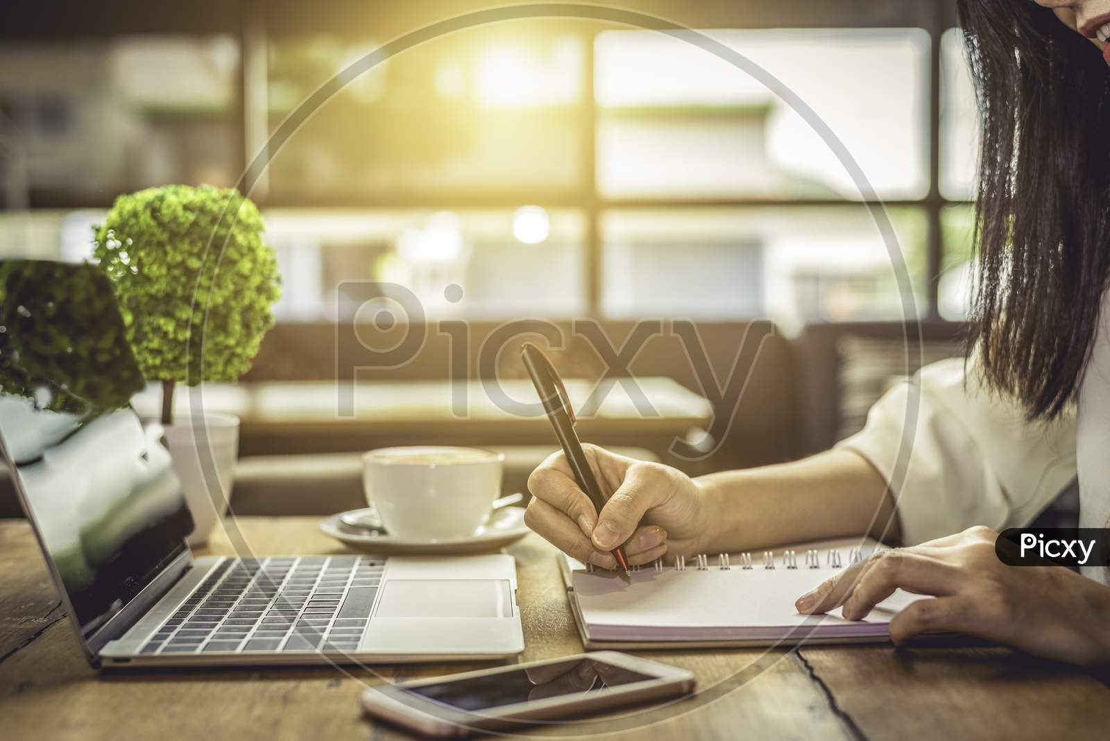 Close Up Of Woman Hand Writing Business Project Planning In Notebook With Laptop And Smart Phone At Coffee Shop. People And Technology Concept. Freelance And Lifestyle Theme. Entrepreneur And Working