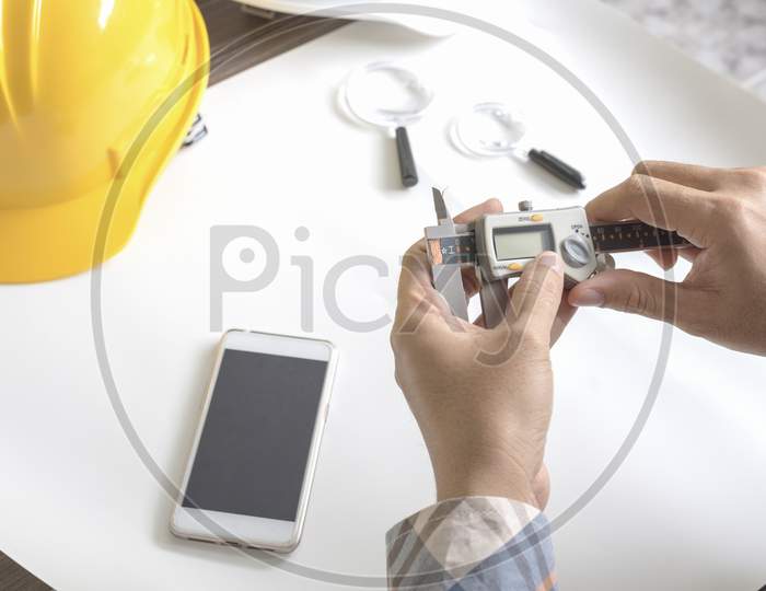 Construction Engineer Measuring With Vernier Caliper. Business And Technology Concept. Safety Helmet And Drawing Paper Elements. Civil And Drawing Sketch Theme. Smart Phone Element