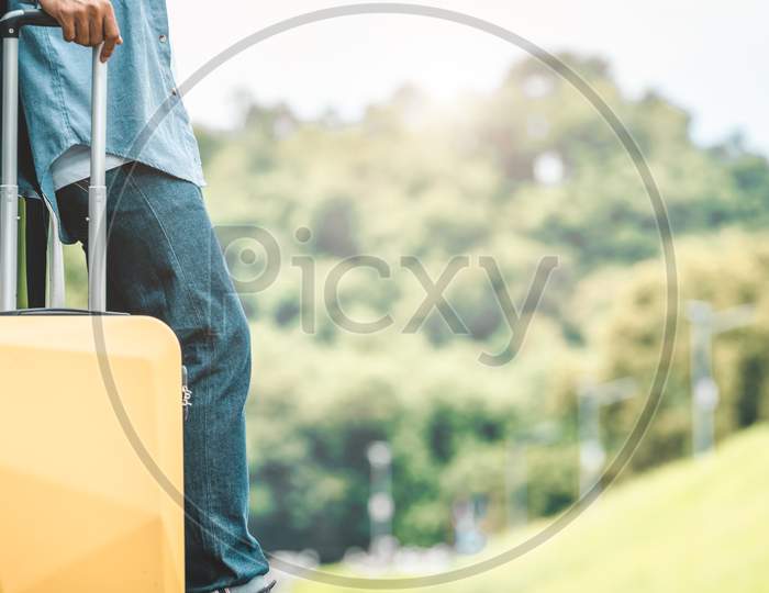 Closeup Of Man Legs Resting During Travel On Road Trip With Yellow Suitcase On Summer Nature Background. Transportation And People Lifestyles Concept. Tourist Standing Outdoors On Vacation Tour
