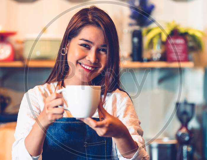 Asian Female Barista Making Cup Of Coffee. Young Woman Holding White Coffee Cup While Standing Behind Cafe Counter Bar In Restaurant Background. People Lifestyles And Business Occupation Concept.