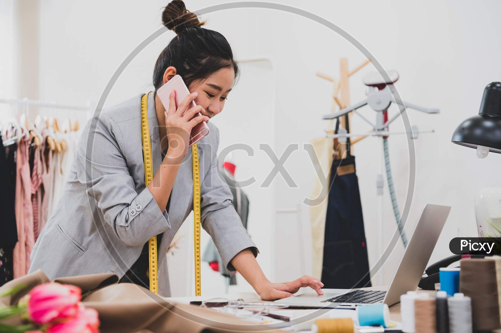 Attractive Asian Female Fashion Designer Working In Clothing Shop Studio And Using Mobile Phone To Contact Customer. Woman Receiving Order From Incoming Call. Tailor Sewing. People Lifestyle Concept