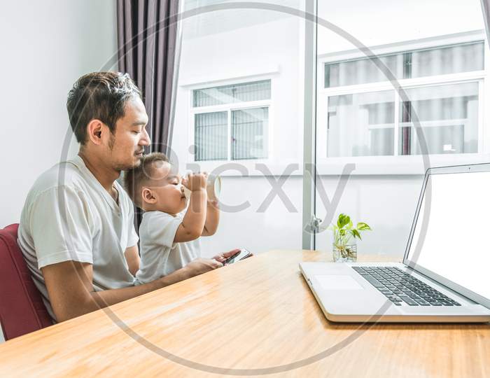 Asian Father And Son Using Smart Phone And Laptop Together In Home Background. Son Drinking Milk. Technology And People Concept. Lifestyles And Happy Family Theme. Internet And Communication Theme