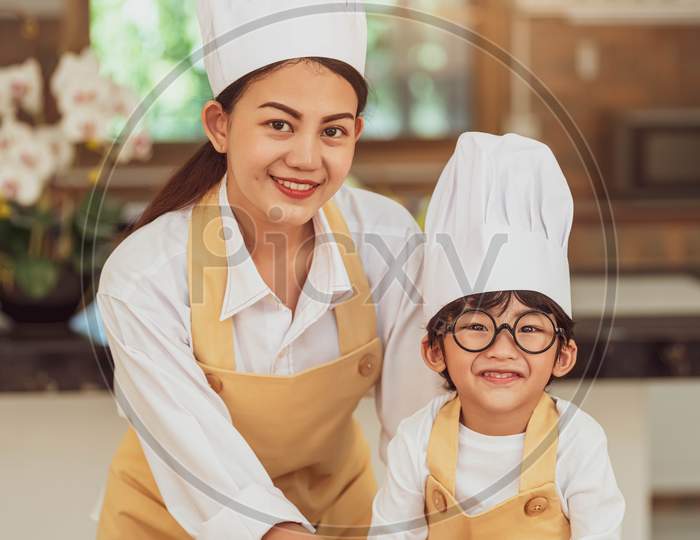 Portrait Cute Little Asian Boy Chef With Eyeglasses And His Mother Looking To Camera In Home Cooking Kitchen Happily And Funny. People Lifestyles And Family. Homemade Food Ingredient Making Concept