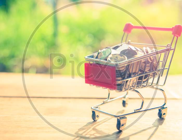 Shopping Cart On Wooden Table With Coins In Outdoor Nature Background. Money Saving And Income Concept Idea. Selective Focus On Coin. Pastel Tone