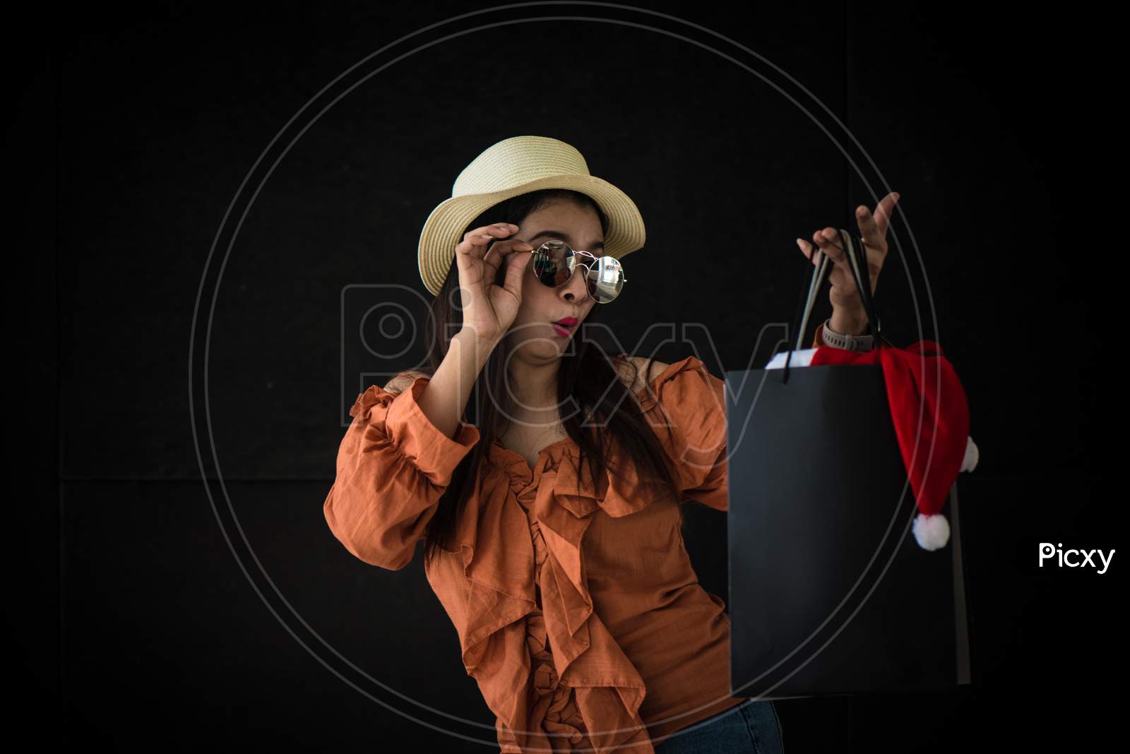 Asian Shopping Woman Surprised With Black Friday Shopping Bag And Santa Claus Hat Inside On Black Background. Shopaholics And Beauty Fashion Theme.