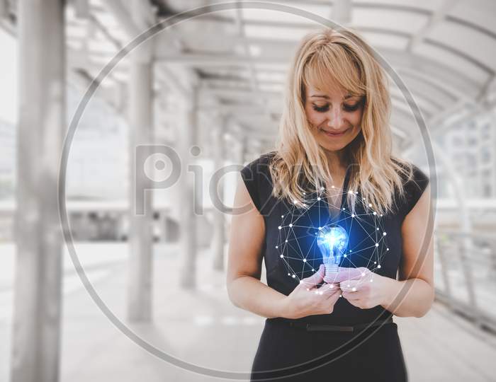Blonde Hair Woman Holding And Looking At Blue Light Bulb With Bright And Shine Line Dot Illustration Effect. Future Technology And Lifestyle Concept. Beauty And Object Concept. City And Urban Theme.