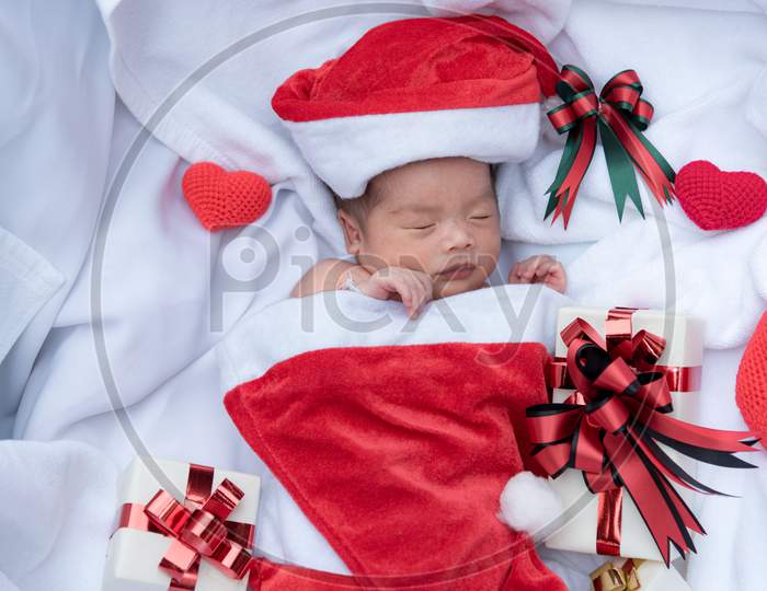 Sleeping Newborn Baby Face In Christmas Hat With Gift Box From Santa Claus And Yarn Heart On White Soft Towel. Cute Infant Lifestyle And Innocent Happy Baby Lying In Cold Snow Season. New Year Winter