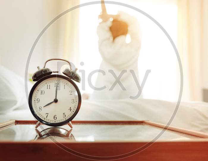 Back View Of Woman Stretching In Morning After Waking Up On Bed Near Window With Alarm Clock. Holiday And Relax Concept. Lazy Day And Working Day Concept. Office Woman And Worker In Daily Life Theme