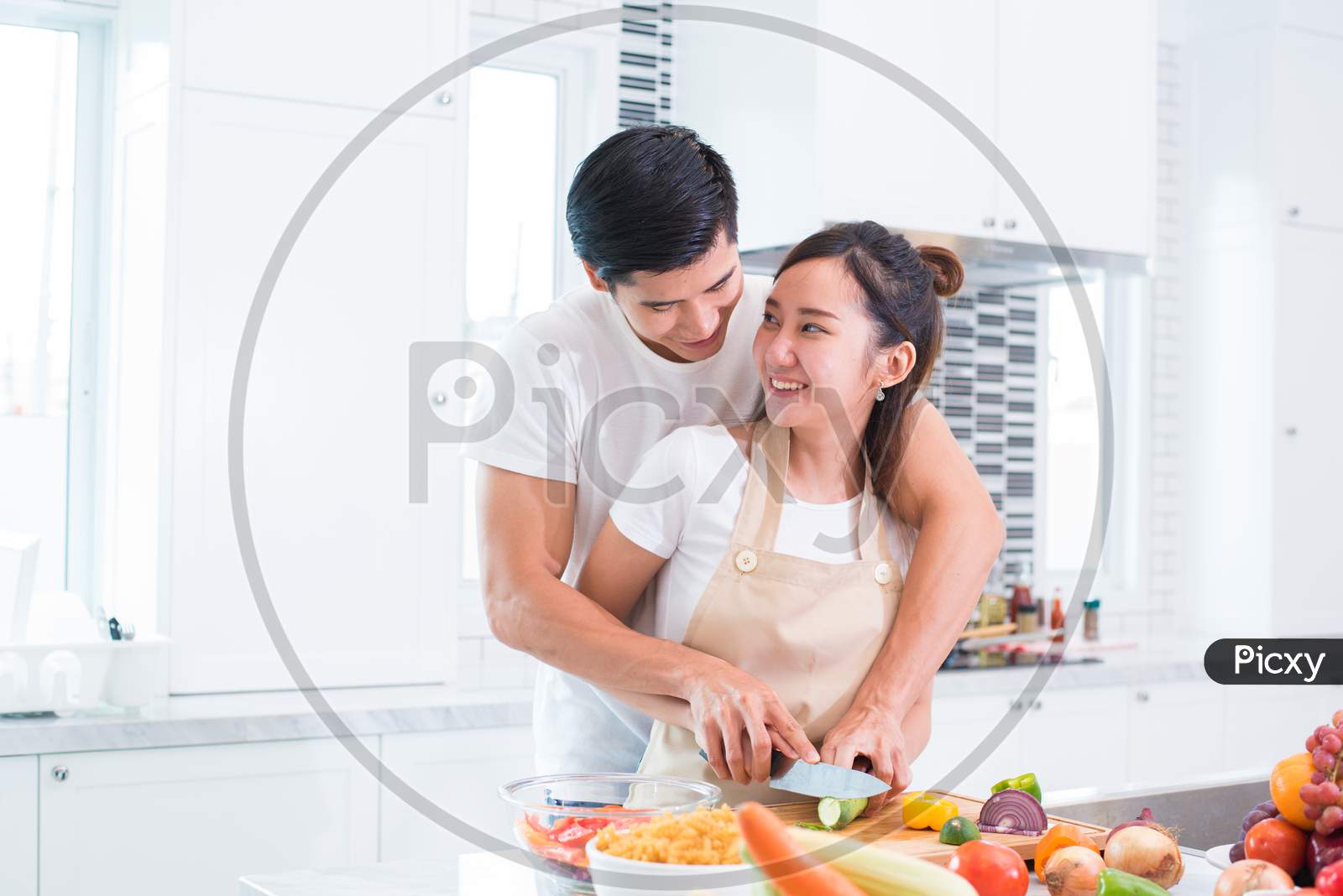 Asian Lovers Or Couple Cooking And Slicing Vegetable In Kitchen Room. Man And Woman Looking Each Other In Home. Holiday And Honeymoon Concept. Valentine Day And Wedding Theme