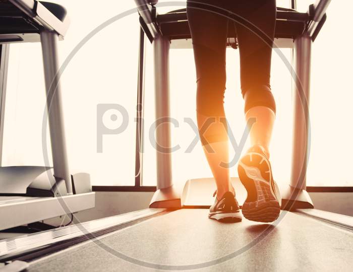 Lower Body At Legs Part Of Fitness Girl Running On Running Machine Or Treadmill In Fitness Gym With Sun Ray. Warm Tone. Healthy And Exercise Activity Concept. Workout And  Strength Training Theme.