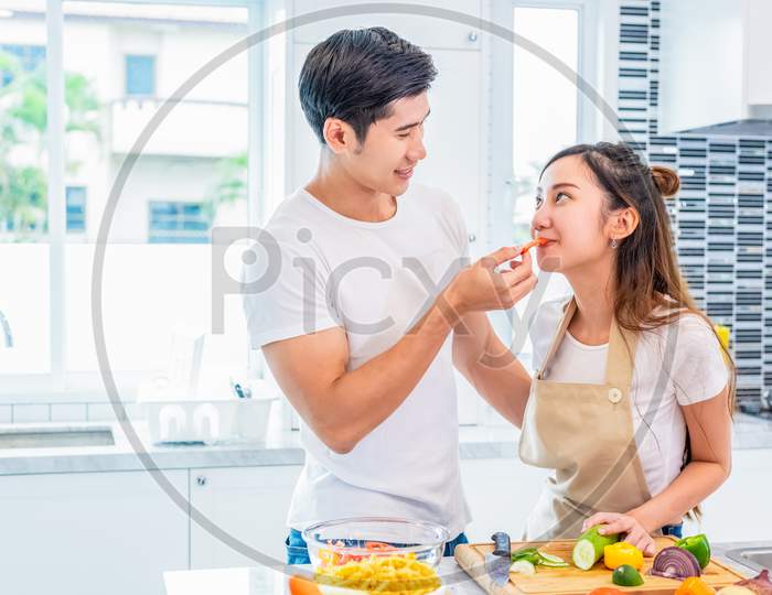 Asian Boyfriend Feeding Fruit And Vegetable To Girlfriend To Each Other During Preparing For Dinner In Home Kitchen. Couple And Family Concept. Honeymoon And Holidays. Man And Woman Cooking Happily