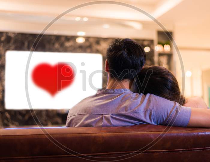 Asian Young Lovers Watching Television On Sofa. Couples And Relax Concept. Holiday And Vacation Concept. Night Dating And Honeymoon Theme. Back View. Red Heart In White Screen Television