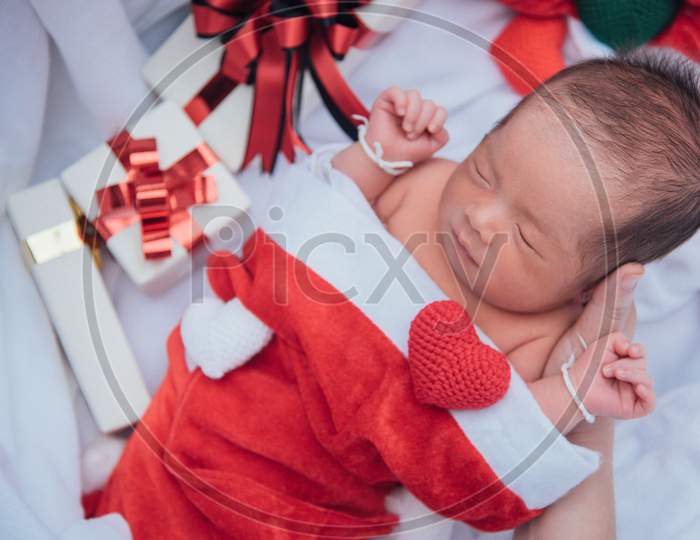Sleeping Newborn Baby On Mother Hand In Christmas Hat With Gift Box From Santa Claus And Yarn Heart On White Soft Towel. Cute Infant Lifestyle And Innocent Lying In Cold Snow Season. New Year Winter