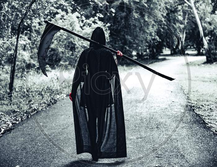 Back View Of Demon Witch With Reaper And Red Blood On Hands Walking Along The Road Background. Horror And Ghost Concept. Halloween Day And Scary Scene Theme. Killer And Death Theme.