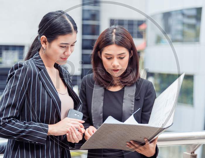 Two Young Businesswomen Looking Into Document File Folder For Analyzing Profit Circulation Or Sale Break Even Point After Marketing. Business Teamwork Employees Of Lifestyle Working Women Concept.