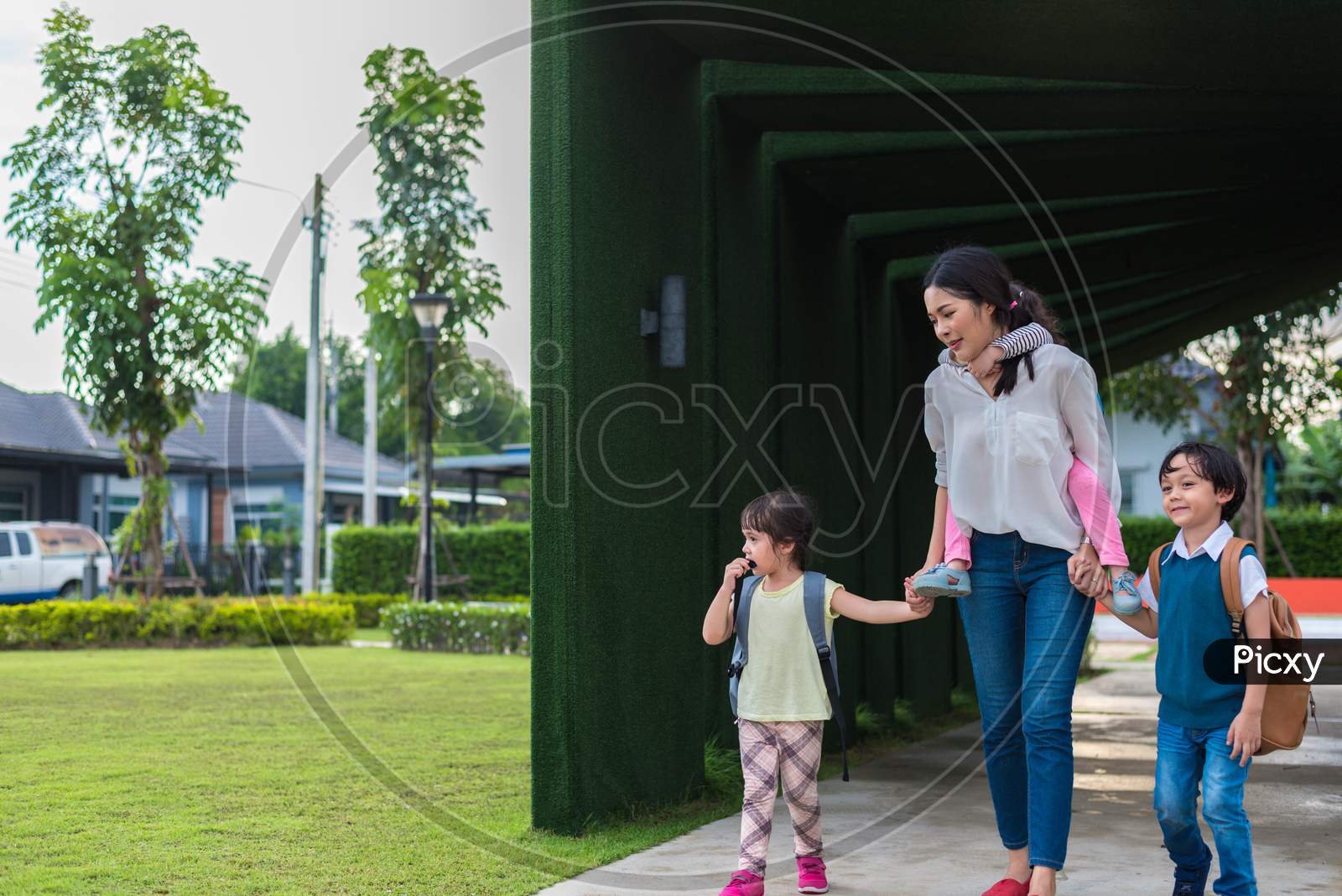 Single Mom Carrying And Playing With Her Children In Garden With Green Wall Background. People And Lifestyles Concept. Happy Family And Home Sweet Home Theme. Outdoors And Nature Theme.