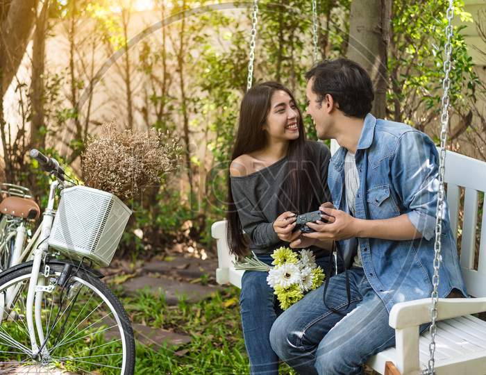 Asian Couple Having Eye Contact As Romantic Moment At Bench In Natural Park With Bicycle. People And Lifestyles Concept For Valentines Day And Wedding Ceremony. Love At First Sight Theme.