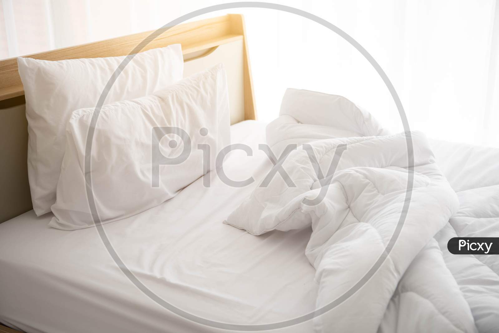 White Unmade Bed In The Morning With Sunshine With White Curtain Background In Elegant Home. Interior Design And Luxury Furniture Concept. Indoors Lifestyle And Lazy Activity Theme.
