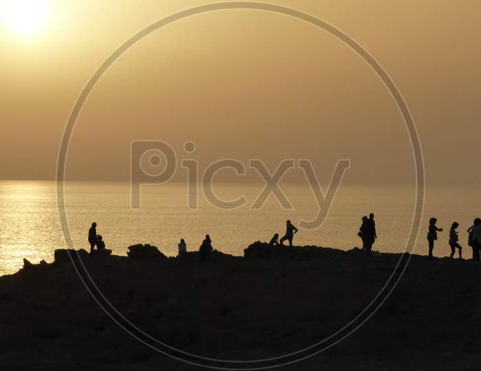 People Watch The Sunset Near The Ocean. A Darkened Image.