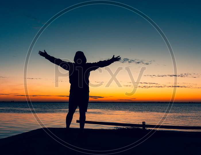 Shocking Sunset At The Edge Of The Lagoon. The Sun Sets On The Horizon Painting Everything In Golden Colors. Silhouette Of A Hooded Man With Outstretched Arms.