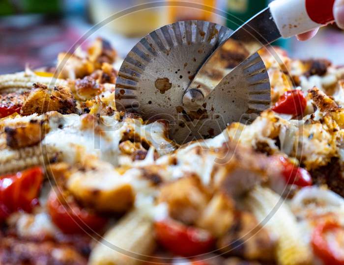 Macro View Of A Circular Pizza Cutter Cutting Through A Pizza Filled With Cheese, Baby Corn, Tomatoes, Chillies And More On A Vegetarian Pizza