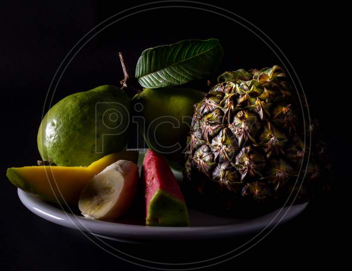 Whole And Cut Varied Fruits On A Plate. Neutral Black Background With Pictorial Lighting. Vegetarian Or Vegan Breakfast Concept.