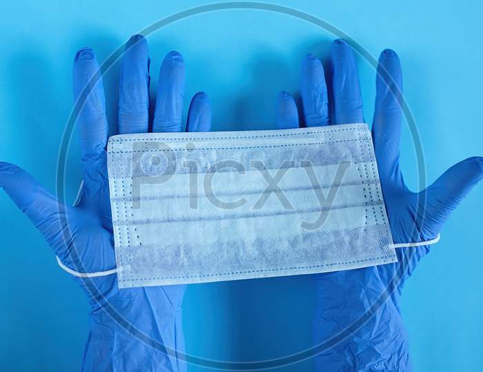 Two Hands Of A Doctor In Medical Gloves Holds Medical Surgical Mask. Ready For Covering The Mouth And Nose. Top View, Close Up, On Blue Background