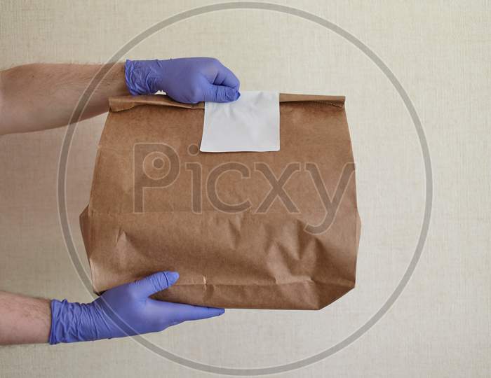 Male Hands In Blue Latex Glove Gives Paper Bag To Female Hands. Closeup Of Male Hands In Gloves Hold Brown Craft Paper Bag With Blank White Sticker. Delivery Service Concept. Copy Space Area.