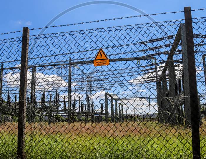 Sign Showing The Words High Voltage In German Language At The Fence Of A Big Substation Distributing Electric Energy With Lots Power Lines On A Sunny Day