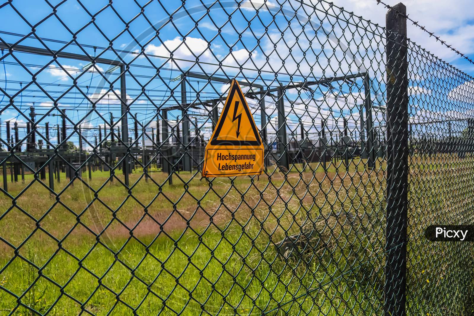 Sign Showing The Words High Voltage In German Language At The Fence Of A Big Substation Distributing Electric Energy With Lots Power Lines On A Sunny Day