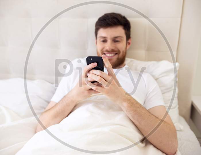 Man Chatting On Phone In Bed. Morning At Home. Smilling Man Using Phone In Bed. Quarantine, Home Work, Medical Care.