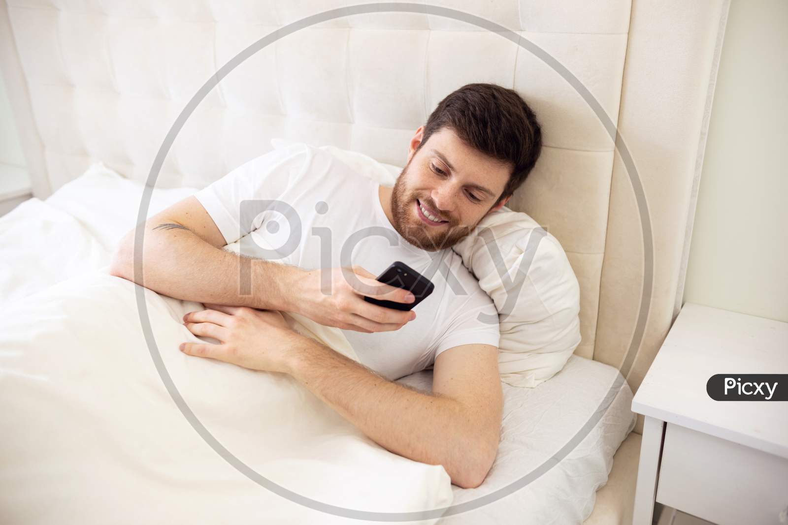 Man Chatting On Phone In Bed. Morning At Home. Smilling Man Using Phone In Bed. Quarantine, Home Work, Medical Care.