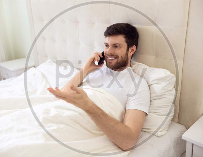 Man Talking On Phone In Bed. Morning At Home. Smilling Man Using Phone In Bed. Quarantine, Home Work, Medical Care.