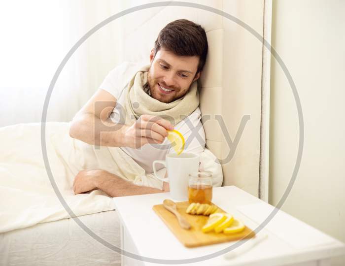 Sick Man In Bed Adding Lemon Into Tee Smilling. Man Ill In Bed Self Healing. Self Treatment At Home. Man Drinking Hot Tea In Bed.