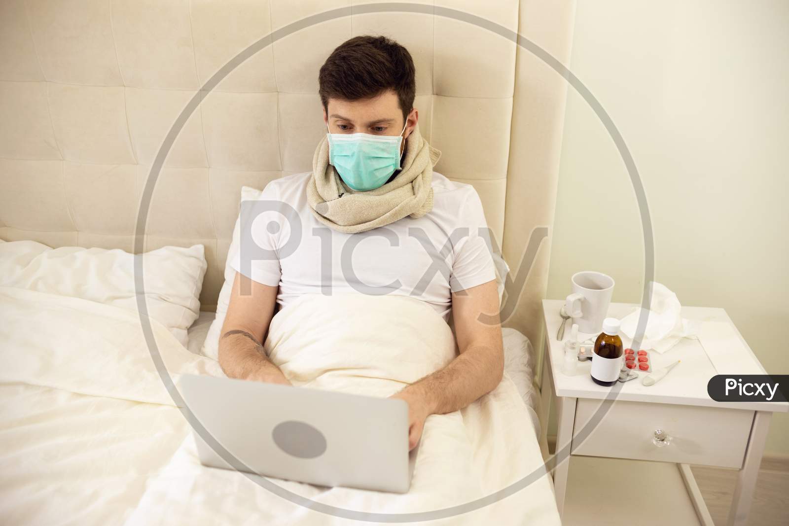 Sick Man Working In Bed Wearing Medical Mask. Man Working At Home. Man Using Laptop In Bed. Man Ill At Home. Quarantine, Home Work, Medical Care.