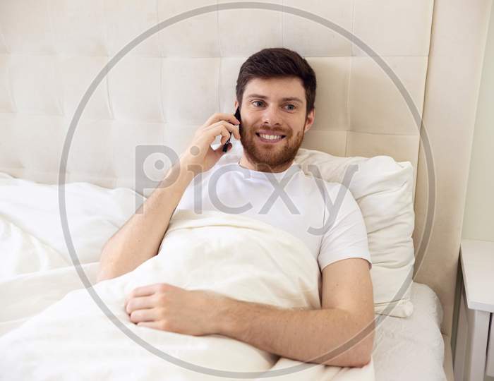 Man Talking On Phone In Bed. Morning At Home. Smilling Man Using Phone In Bed. Quarantine, Home Work, Medical Care.