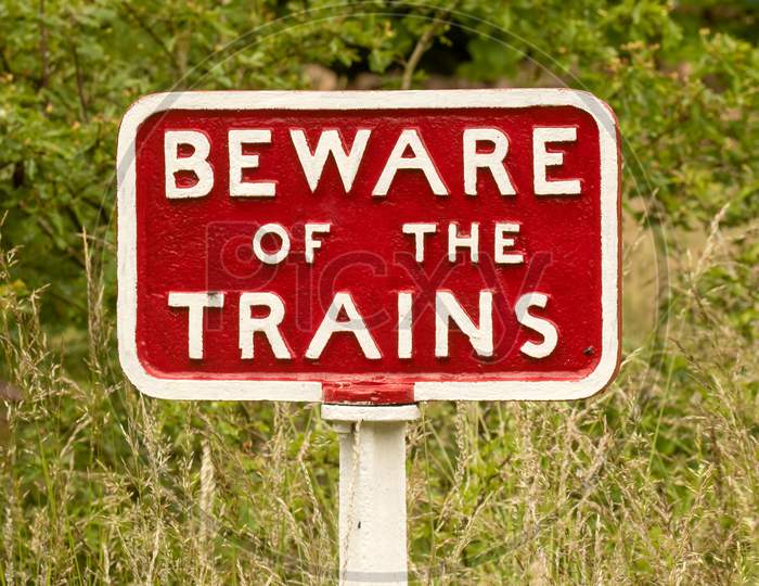 Beware Of The Trains Sign, Cast Iron - Red With White Lettering, Against A Grassy Background