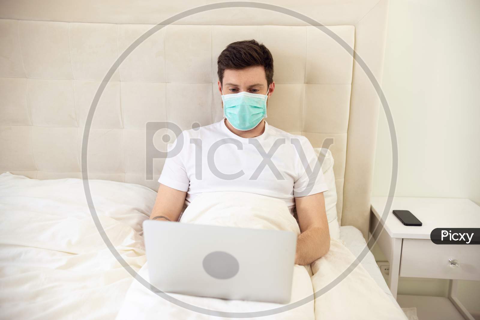 Man Working In Bed Wearing Medical Mask. Man Working At Home. Man Using Laptop In Bed. Quarantine, Home Work, Medical Care.