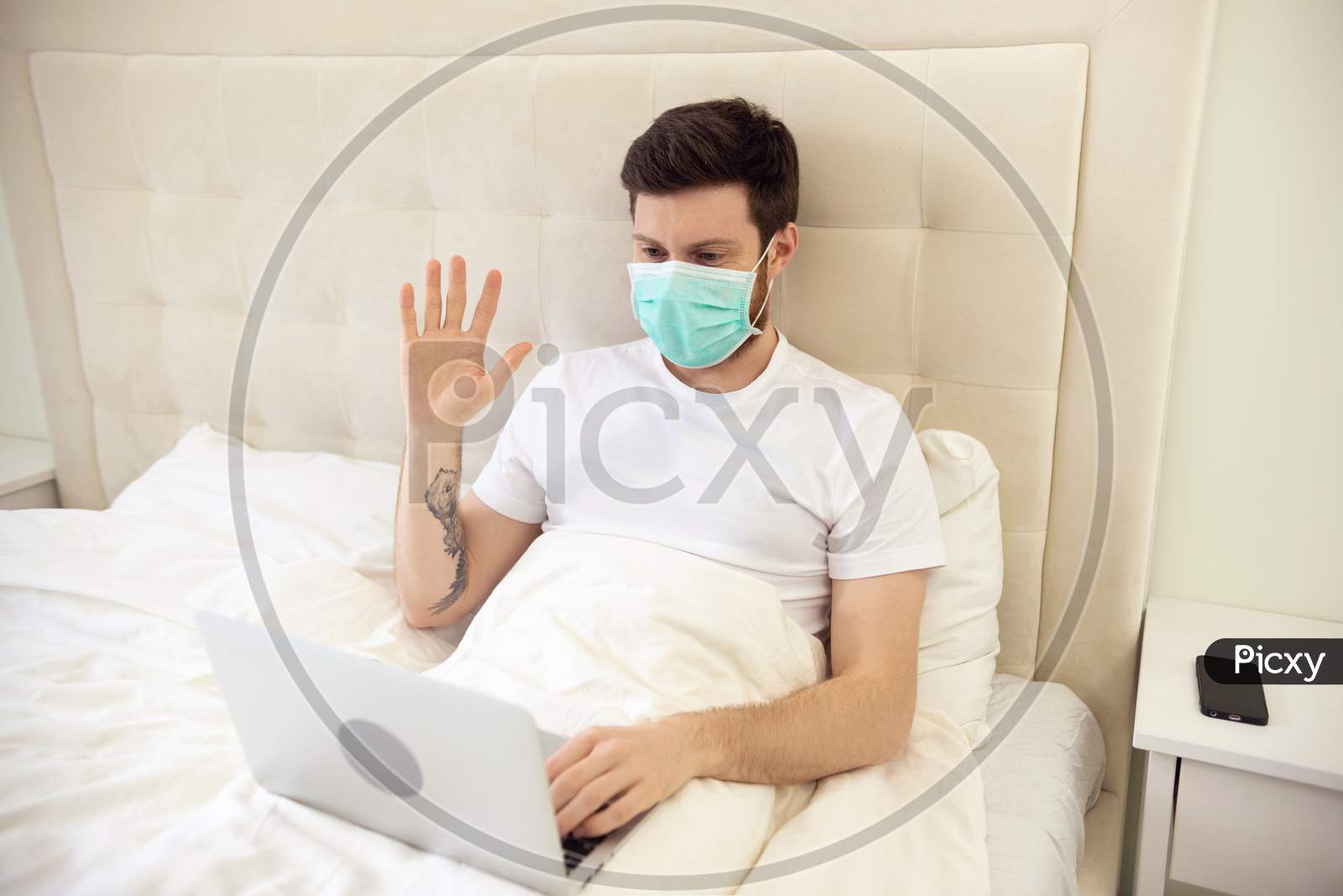 Man Having Video Call Video Conversation In Bed Wearing Medical Mask. Man Working At Home. Man Using Laptop In Bed. Man Ill. Quarantine, Home Work, Medical Care.