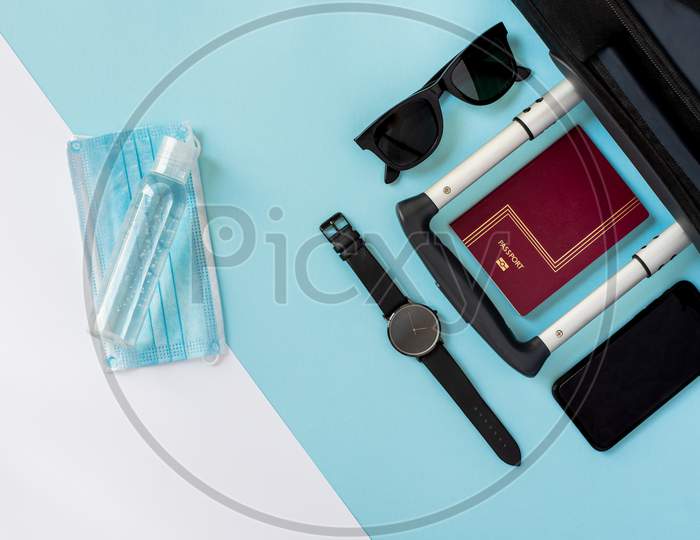 Accessories Of A Traveller On The Summer Holidays 2020 With The Coronavirus. Black Suitcase, Face Mask, Hydroalcoholic Gel, Clock, Smartphone, Sunglasses And Passport