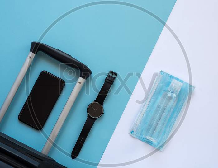 Accessories Of A Traveller On The Summer Holidays 2020 With The Coronavirus. Black Suitcase, Face Mask, Hydroalcoholic Gel, Clock And Smartphone.