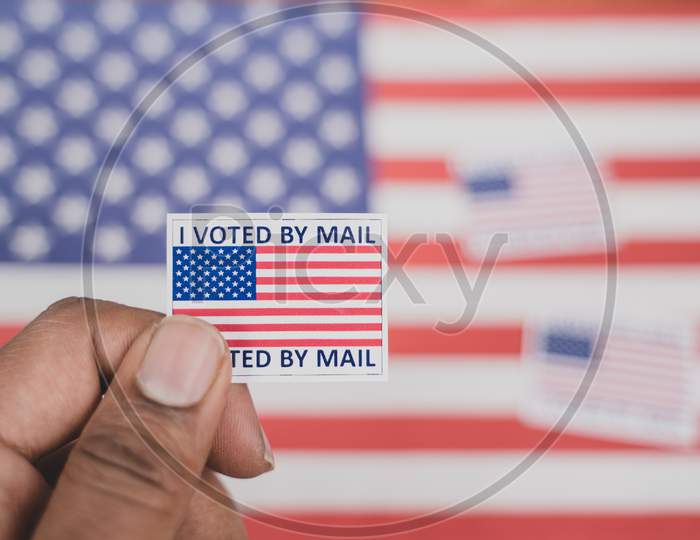 Holding I Voted My Mail Sticker In Hands With Us Flag As Background - Concept Of Voted Through Mail During Election
