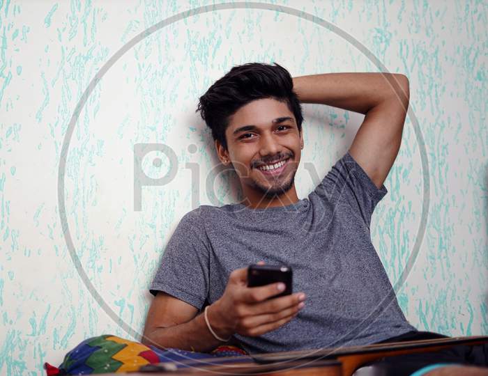 Young Indian Boy Smiling While Looking Into The Camera, With Phone In One Hand And The Other Behind His Head.