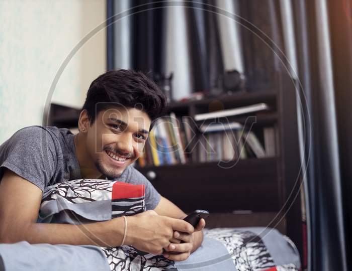Young Indian Boy Smiling Into The Camera, While Using His Phone Lying On His Bed.